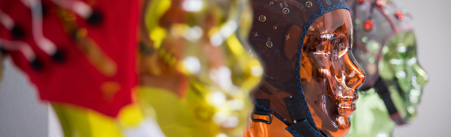 Photo of various EEG masks with stands in the shape of heads on a shelf. The focus is on an orange glass head in the center. __ Various EEG masks with stands in the shape of a face are lined up on a shelf. The focus is on the orange glass head in the middle.