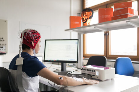 Foto eines Probanden mit EEG Haube und Neurorezeptor an Ellenbogen vor einem Messbildschirm.