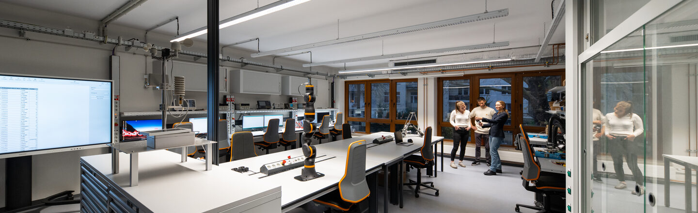 Man sieht ein modernes Labor mit mehreren Arbeitsplätzen, ausgestattet mit Computern, Monitoren und technischem Equipment. In der Mitte des Raums befinden sich Werkbänke mit ergonomischen Stühlen, Steckdosenleisten und einem Roboterarm. Im Hintergrund stehen drei Personen, die sich unterhalten.