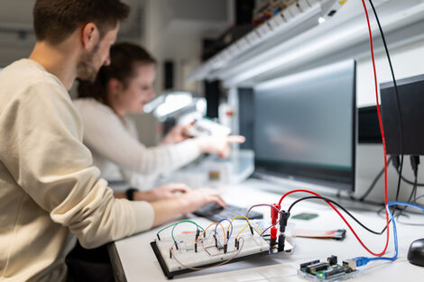Zwei Personen arbeiten in einem Labor an einer elektronischen Schaltung. Im Vordergrund liegt ein Steckbrett mit Kabeln und Bauteilen auf dem Tisch. Die Arbeitsstation ist mit Messgeräten, Werkzeugen und Beleuchtung ausgestattet.