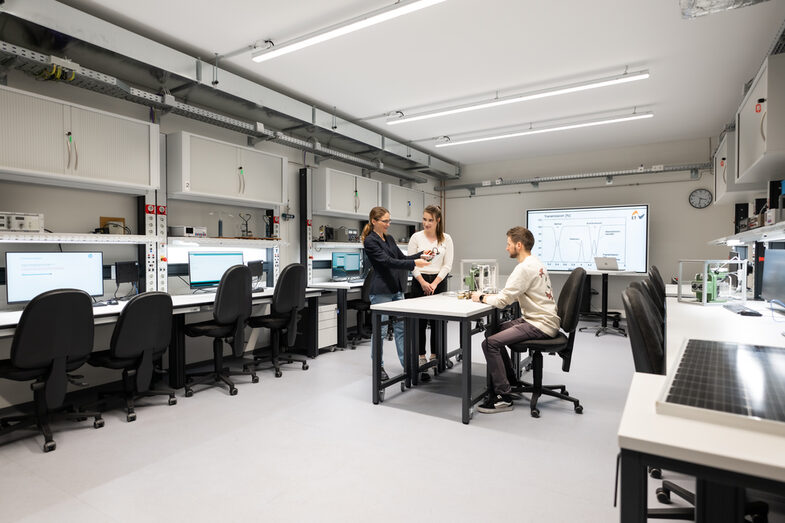 Man sieht ein modernes Labor mit mehreren Arbeitsplätzen, ausgestattet mit Computern, Monitoren und technischem Equipment. In der Mitte des Raums sieht man eine Person die an einem Tischt sitzt und vor ihr zwei Personen stehen, die sich miteinander unterhalten.