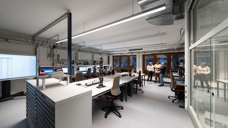 You can see a modern laboratory with several workstations equipped with computers, monitors and technical equipment. In the middle of the room are workbenches with ergonomic chairs, power strips and a robotic arm. In the background are three people having a conversation.