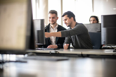 Photo of a lecturer next to a student at a PC. The lecturer is showing the student something on the screen. In the background is another student at a computer.