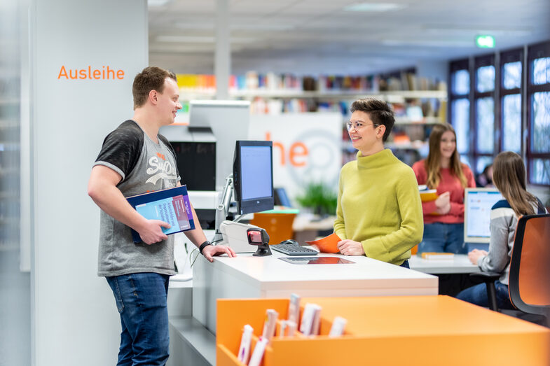 Photo of a member of staff behind the library counter, she is in conversation with the student standing opposite her, who is holding books. In the background, another student is talking to a member of staff.