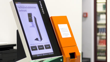 Foto eines Selbstverbuchungsgeräts in der Bibliothek, daneben ein Stapel Bücher auf einem Buchwagen__Photo of a self-service check-out machine in the library with a stack of books on a book cart next to it