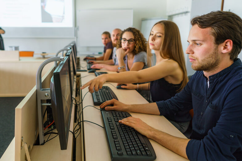 Foto von fünf Studierenden in einer Reihe an PCs. Eine Studentin zeigt den anderen etwas auf dem Bildschirm ihres Sitznachbars.__Reihe an fünf PCs und schauen auf die Bildschirme.__Five students sit in a row at five PCs. One student is showing something to the others on the screen of the student sitting next to her.