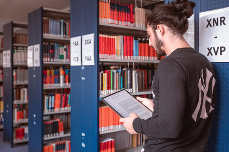 Foot of a young man standing in a library holding a tablet.