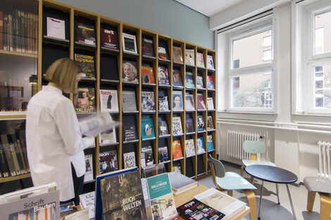 Photo of a female student standing in front of a library journal shelf. The room also features chairs, a table with books and a display cabinet with historic books__Photo of a female student standing in front of a library journal shelf. The room also features chairs, a table with books and a display cabinet with historic books