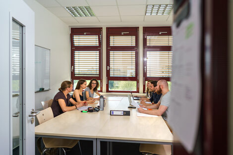 Foto in einen Raum. Sechs Studierende an einem Gruppentisch tauschen sich aus. Auf dem Tisch liegen Arbeitsmaterialien.__View into a room. Six students at a group table exchange ideas. Working materials are on the table.