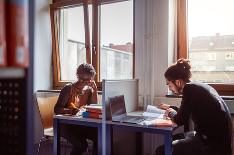 Photo of a young woman and a young man studying with books and laptops at two tables in the library.