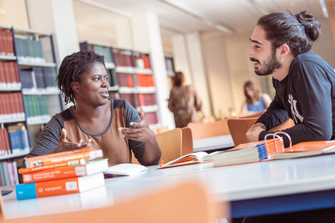 Photo of a young woman and a young man sitting at a table in the library and talking. There are some books on the table, in the background you can see bookshelves and two other people studying.