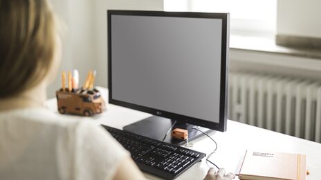 Foto einer Frau am Arbeitsplatz, sie schaut auf einen PC-Monitor. Auf dem Schreibtisch ist eine Stiftebox und ein Notizbuch. __ Woman at work, looking at calculator. On the desk is a pen box and a notebook.