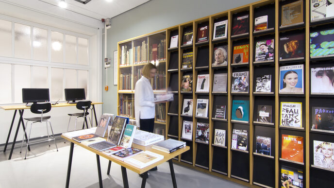 Foto einer Studierenden vor dem Zeitschriftenregal in der Bibliothek. In dem Raum befinden sich auch ein Tisch mit Büchern, eine Vitrine mit historischen Büchern und zwei Computer-Arbeitsplätze__Photo of a female student standing in front of a library journal shelf. The room also features a table with books, a display cabinet with historic books and two computer desks