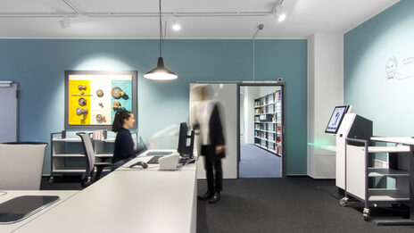 Foto einer Studierenden, die einer Mitarbeiterin der Bibliothek an der Ausleihtheke ein Buch übergibt__Photo of a female student handing a book to a female member of library staff at the service counter