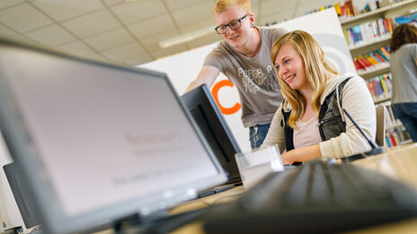 Foto von einem Studenten, der einer Studentin etwas an einem Coputerbildschirm an Computerarbeitsplätzen in der Bibliothek zeigt.__A male student shows something to a female student on a computer screen at computer workstations in the library.