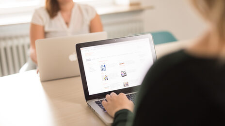 Photo of a young woman at the laptop, sitting across from her at the table, another woman with a laptop. __ Young woman at the laptop, sitting across from her at the table, another woman with a laptop.