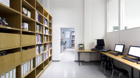 Foto eines Raums in der Bibliothek, in dem sich ein Bücherregal, ein Scanner und mehrere Computer-Arbeitsplätze befinden__Photo of a room in the library featuring a book shelf, a scanner and several computer desks