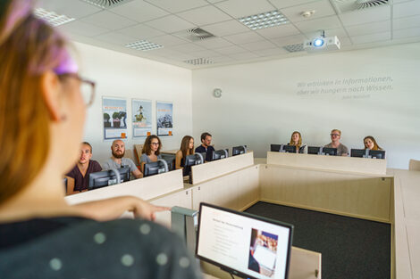 Foto von acht Studierende in einem Seminarraum an PCs. Sie hören einer Frau vorne links im Bild im Anschnitt zu.__Eight students are sitting at PCs in a seminar room listening to a young woman in the front left corner of the picture.