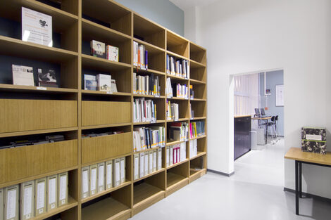 Foto eines Bücherregals in der Bibliothek, im Hintergrund Schließfächer und zwei Computer-Arbeitsplätze__Photo of a library book shelf with lockers and two computer desks ins the background