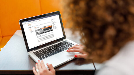 Foto einer jungen am Laptop, im Hintergrund sind orangefarbene Sitzmöbel. __ Young woman at the laptop, in the background there are orange colored chairs.