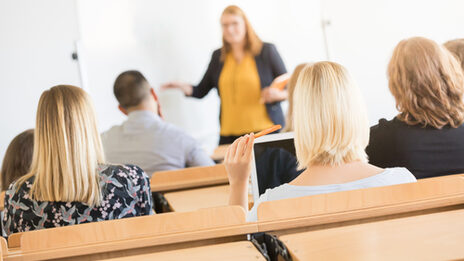 Foto von Hinterköpfen mehrerer Studierender in Sitzreihen im Hörsaal. Vorne ist eine Dozentin unscharf zu erkennen.