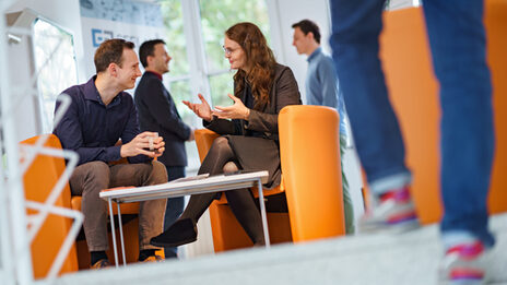 Photo of two people sitting and talking in the hallway, in the background are two other people having a conversation. Another person is coming up the stairs.