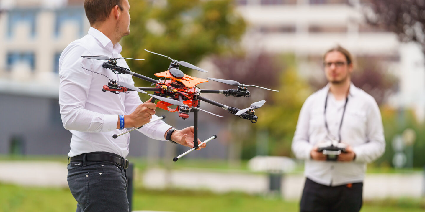 Photo of two IDiAL employees standing opposite each other on a lawn. One of them is holding a drone, the other is holding the controller for controlling the drone.