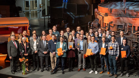 Photo of a group of people. This group is standing on a stage. Some are holding certificates in their hands.