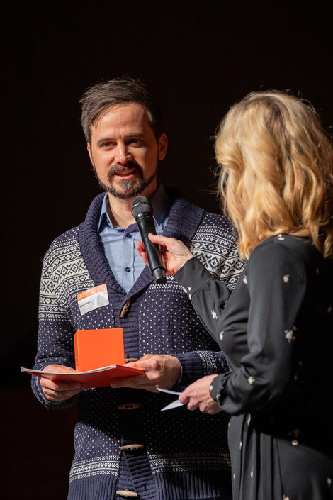 Dr. Robert Höttger nimmt seinen Preis entgegen. Dazu sagt er Worte in ein Mikrofon, das von einer blonden Frau gehalten wird.