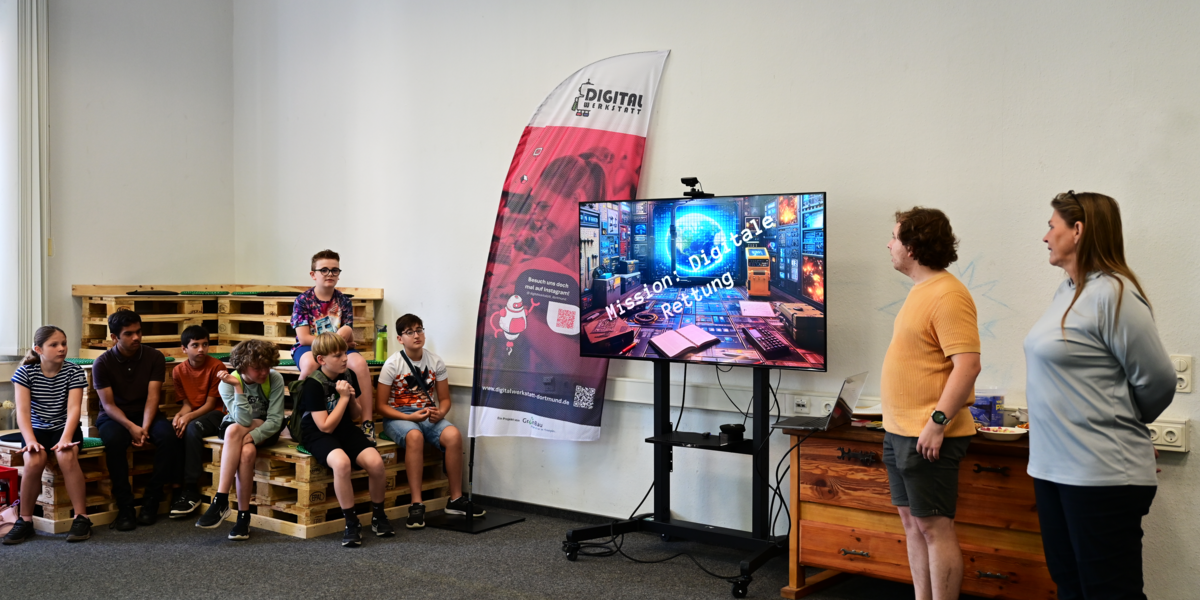 Sieben Schüler im linken Bildbereich hören Sabine Sachweh und Marcel Mitas bei der Beschreibung des Escape Rooms „Digitale Rettung“ zu.