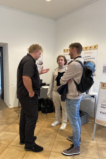 An einem Tisch stehend präsentieren Ansgar Rahmacher, Marcel Mitas und Leon Sachweh Assistenzsystem für die Gesundheits- und Pflegebranche.