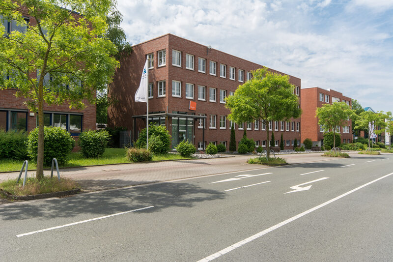 Photo of the location of the Fachhochschule Dortmund in Otto-Hahn-Straße from the outside. It is a red brick building. In the foreground are a road and some trees.