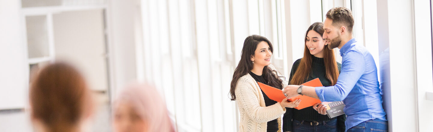 Photo of five international students, two of whom are talking in the foreground and the other three are looking together into an orange folder.