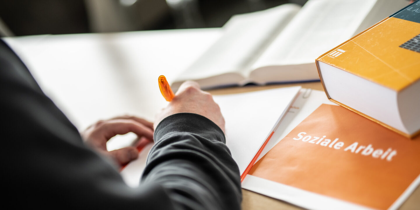 Cropped photo of a person writing something on a pad with an orange pen. Next to it are books and a notebook labeled "social work".