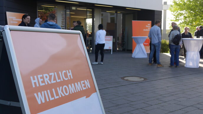 A scene in front of the EFS 44 building. In the foreground you can see a stand with the words "Welcome". In the background, people stand together in small groups.