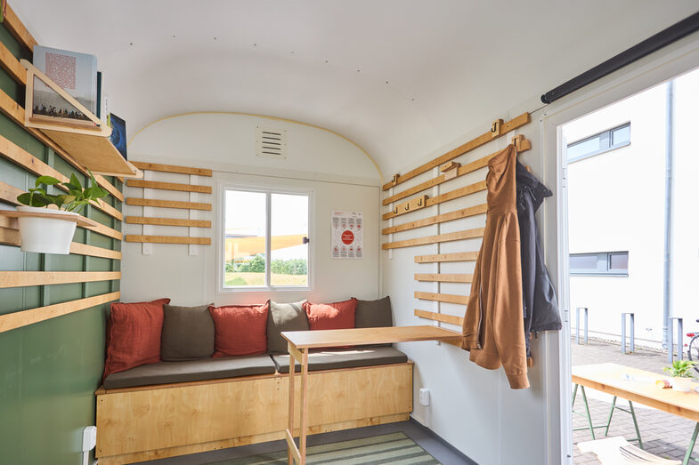 Innenaufnahme eines umgebauten Bauwagens der als mobiles Nachhaltigkeitsbüro dient. Man sieht eine Sitzecke mit mehreren Kissen und einen ausklappbaren Tisch. An der Wand wurden Holz Streifen befestigt, die als Garderobe genutzt werden.