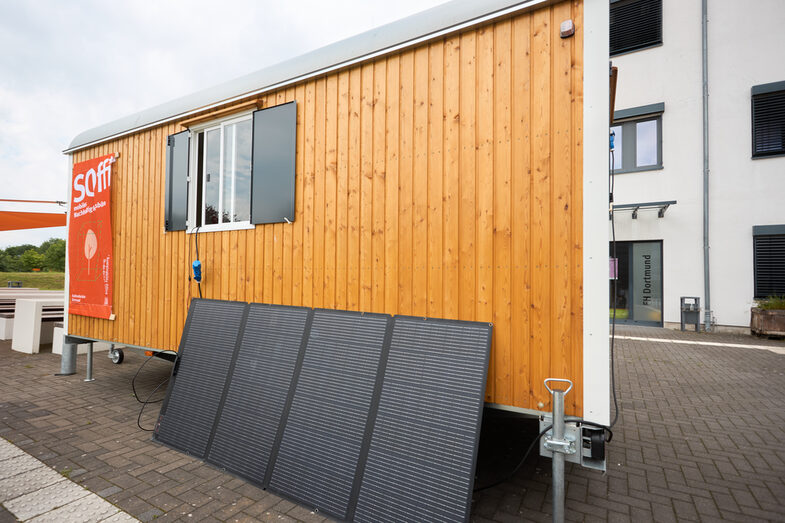 Außenaufnahme eines umgebauten Bauwagens, angelehnt an die Rückseite steht ein Solar Panel. Links am Bauwagen hängt ein Plakat auf dem „Soffi mobiles Nachhaltigkeitsbüro“ steht