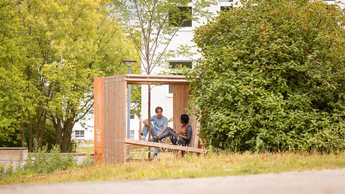 Photo of 2 students sitting and talking in a wooden cube equipped with seating. A tree is planted in the center of the cube.