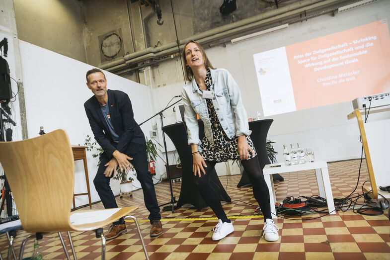 Auf dem Bild stehen zwei Moderatorinnen, die sportliche Bewegungen synchron machen. Im Hintergrund ist eine Leinwand zu erkennen. Die Personen scheinen in ein Publikum zu schauen.