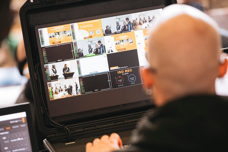 Auf dem Bild sitzt eine Peron mit dem Rücken zur Kamera und schaut auf einen Bildschirm mit mehreren kleinen Kacheln. Es wirkt wie kleine Viedoaufzeichnungen