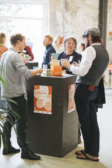 An einem dunklen eckigen Stehtisch stehen in einem hellen Raum drei Personen die sich angeregt unterhalten