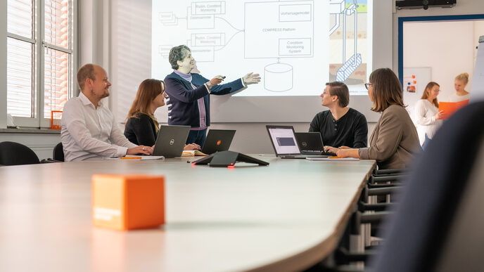 Photo of a meeting situation with 5 people. 4 are sitting at laptops and one woman is presenting standing up. 2 women are recognizable in the background in another room.