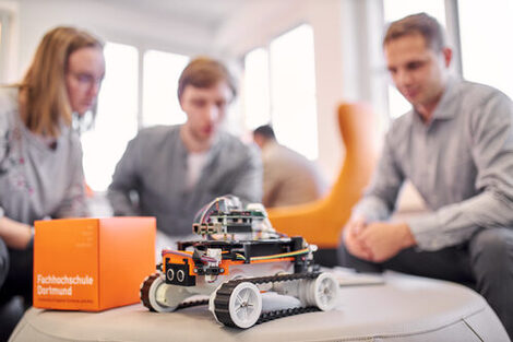 Photo of a 4-wheeled robot standing on a round table together with an orange FH cube. In the background, three people are sitting around it out of focus. They are talking about something.