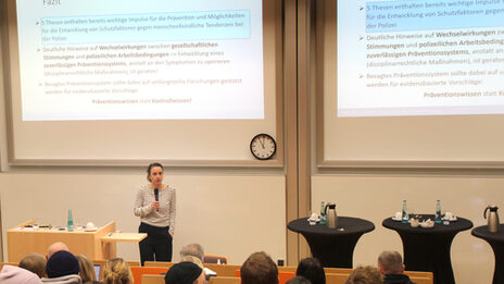 One person stands with their back to a high wall onto which a projector is projecting presentation slides. The person holds a microphone in their hand and speaks to the audience in front of them.