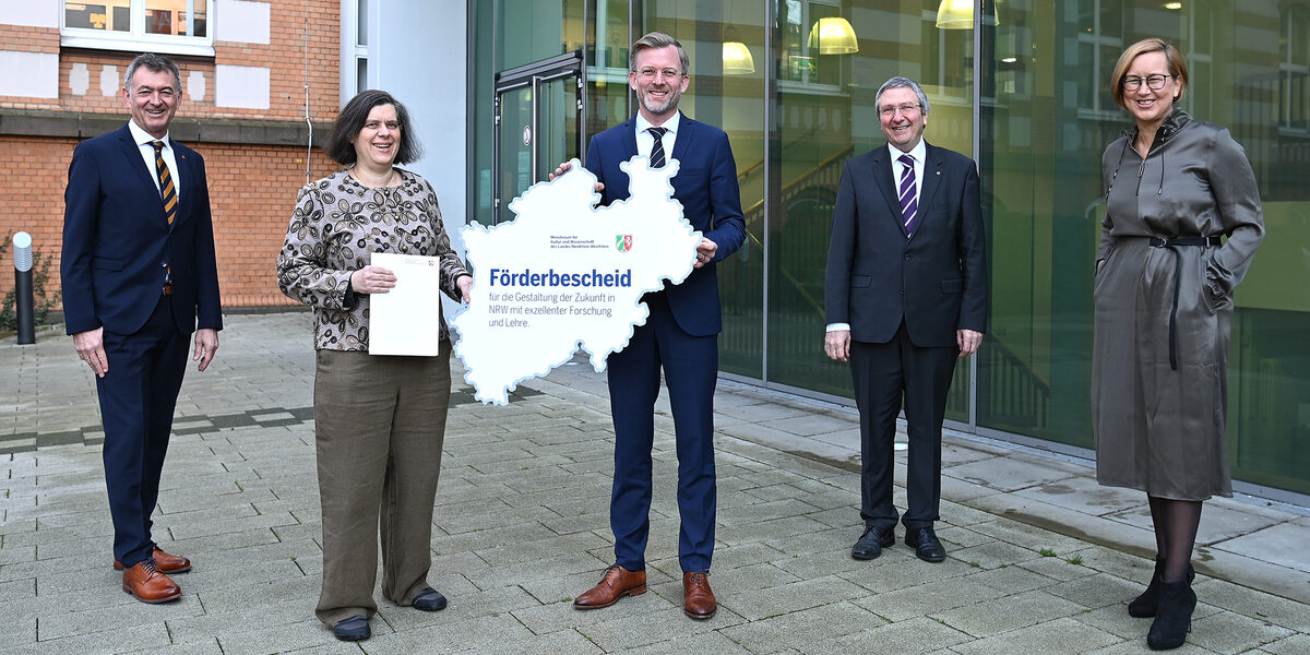 Mehrere Personen stehen mit Abstand vor dem Mensa-Gebäude der FH Dortmund. Zwei von Ihnen halten eine Karte mit den Umrissen von NRW und der Aufschrift "Förderbescheid" in der Hand.