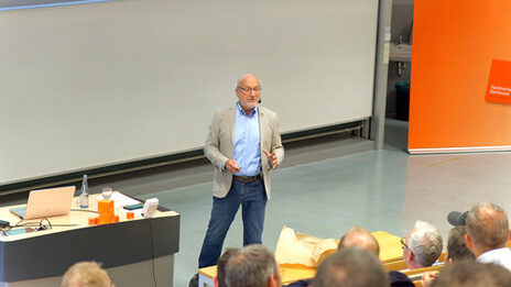 A man stands in a lecture hall and speaks to the people sitting in front of him.