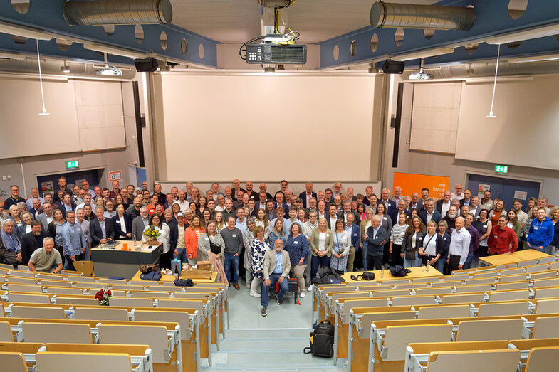 Eine große Gruppe Menschen steht im Hörsaal des Fachbereichs Informatik für ein Gruppenfoto zusammen. Eine Person sitzt vorn auf einem Klavierhocker.