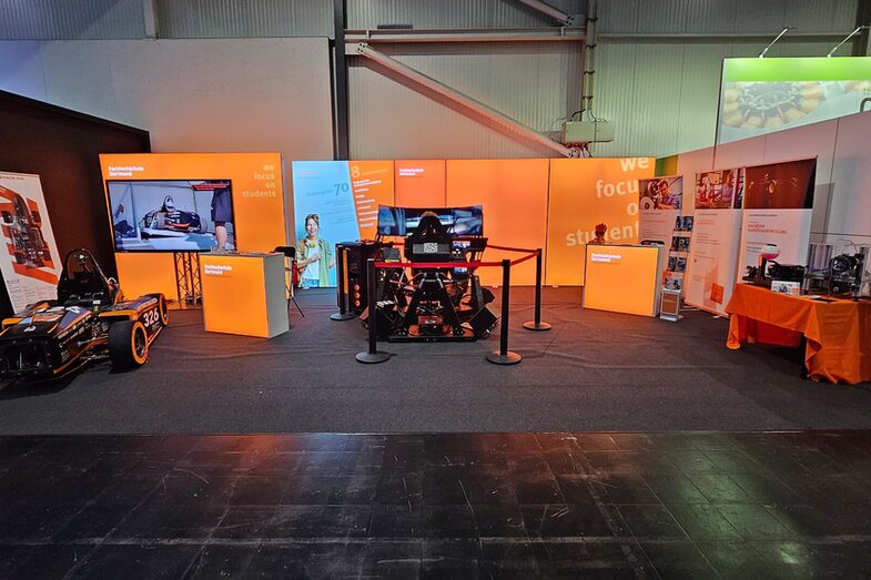 In einer Halle ist ein Messestand aufgebaut, vor einer orangenen Leuchtwand stehen unter anderem ein Rennauto, ein Rennsimulator und ein Tisch mit technischen Elementen.