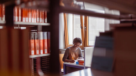 Photo of a student writing something. The camera's gaze passes through a gap in the bookshelf to the student's study area.