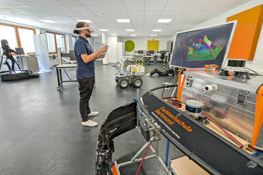 Eine Person mit einer VR-Brille steht in einem Labor. Vor ihr ein Monitor mit bunten Linien.